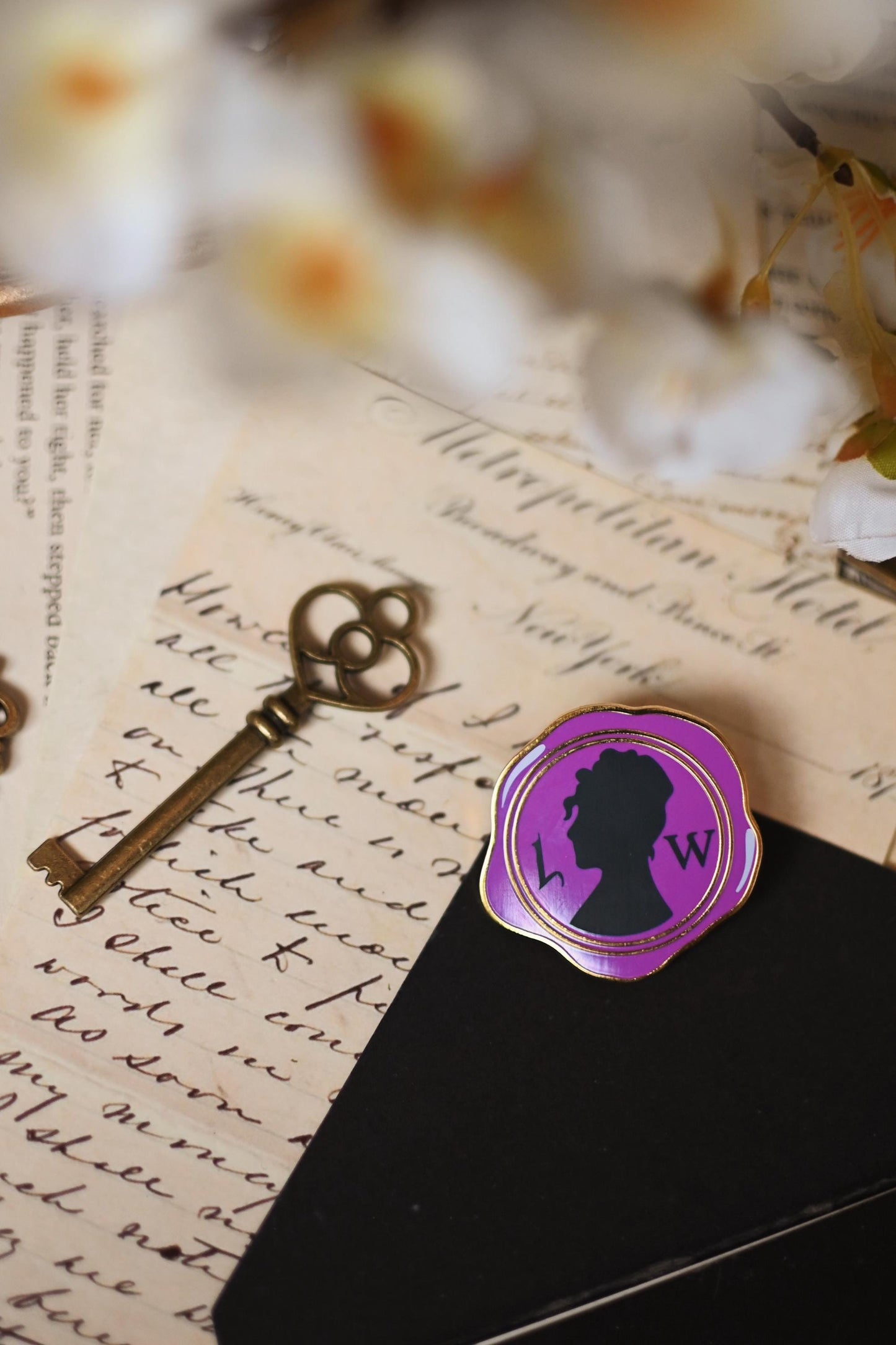 Close up shot of an enamel pin with a purple silhouette of a woman's profile, inspired by Lady Whistledown, placed on a black surface with a vintage background including a key and floral elements.