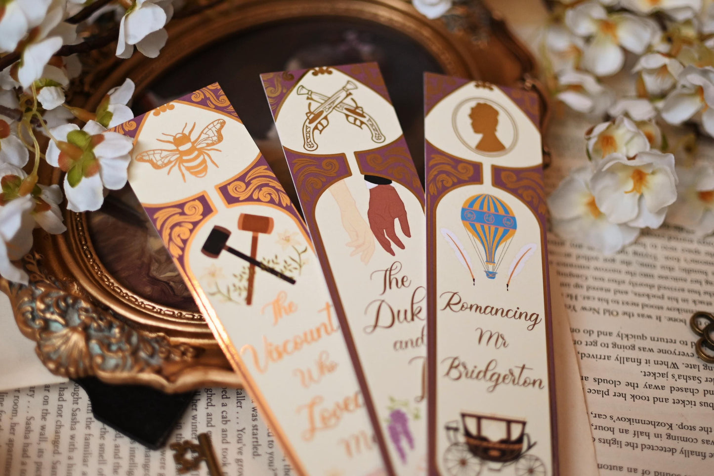 No One Designs Bridgerton-inspired bookmark collection with the three bookmarks fanned out against bookpages and a mirror that raised the bookmarks into the foreground. Each bookmark is inspired by one of Julia Quinn's books and features a gold-foil detailing. Bookmarks inspired by The Duke & I, The Viscount Who Loved Me and Romancing Mr Bridgerton.
