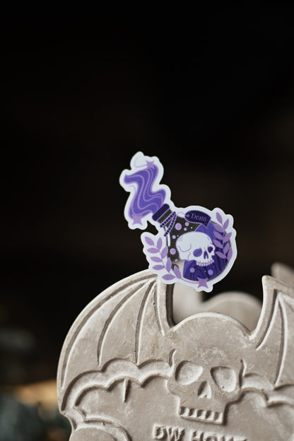 A transparent sticker with a shiny finish designed to resemble a Death Potion Bottle, placed on a surface with written documents and a key in the background.
