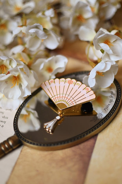 A gold metal enamel pin in the shape of a fan, inspired by the world of Bridgeton, with silk printing and a dangling tassel, displayed on a wooden surface with a key and white flowers in the background.