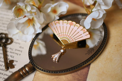 A gold metal enamel pin in the shape of a fan, inspired by the world of Bridgeton, with silk printing and a dangling tassel, displayed on a wooden surface with a key and white flowers in the background.