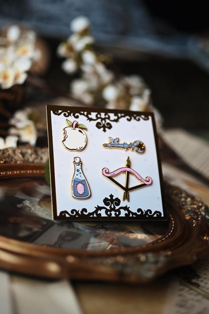 A set of four pins with intricate designs displayed on a card, positioned on a vintage-style surface with floral motifs.