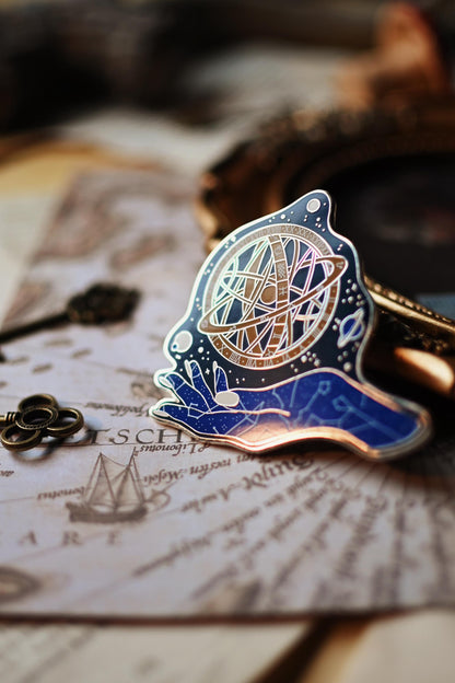 An enamel pin depicting a celestial goddess design with gold, blue, and black colors on a silver metal backing, displayed on a book with written pages.