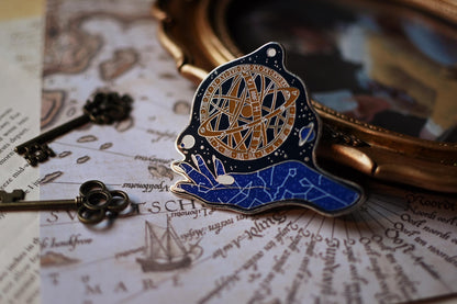 An enamel pin depicting a celestial goddess design with gold, blue, and black colors on a silver metal backing, displayed on a book with written pages.