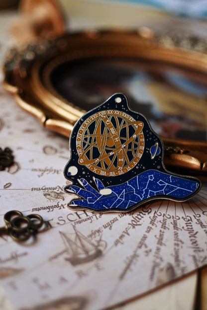 An enamel pin depicting a celestial goddess design with gold, blue, and black colors on a silver metal backing, displayed on a book with written pages.