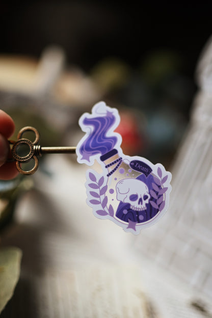 A transparent sticker with a shiny finish designed to resemble a Death Potion Bottle, placed on a surface with written documents and a key in the background.