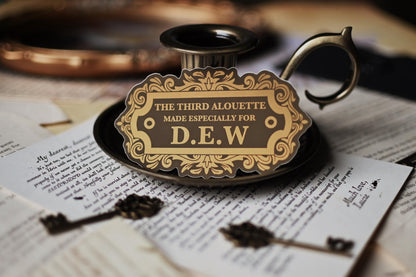 A sticker with vintage design, gold and black in color, with the text 'THE THIRD ALOUETTE especially for D.E.W inspired by Rebecca Ross' Devine Rivals.
