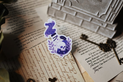 A transparent sticker with a shiny finish designed to resemble a Death Potion Bottle, placed on a surface with written documents and a key in the background.