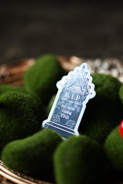 A silver foil decorative sticker with the text 'DEATH BY TBR' on a background that includes green moss and a wooden surface.