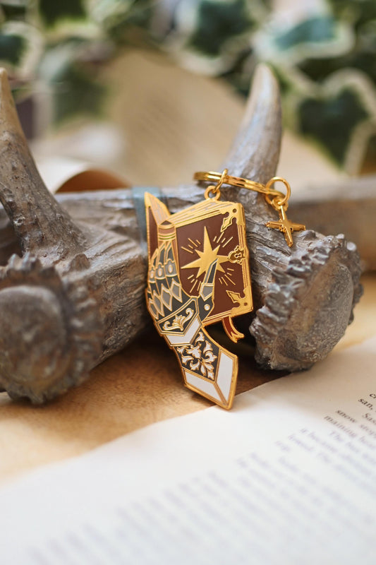A gold metal keychain designed as a gauntlet with a star detail, placed on a wooden surface with a foil backing card.
