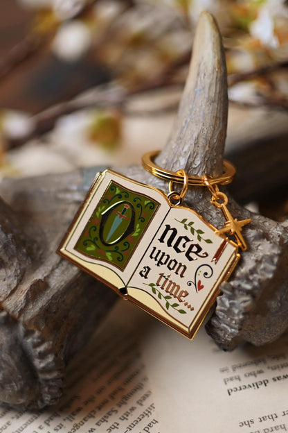 An enamel keychain with a gold metal frame and 'Once Upon a Time' text, displayed on a naturalistic setting with a leafy background.
