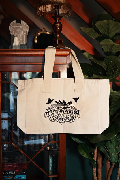 Front on photo of The Crow Club tote bag hanging from a hook of a bookcase showing off the delicate detailing of the logo. 