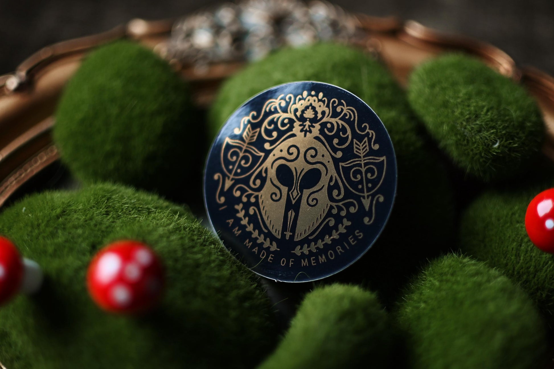 The gloss version of the song of achillies inspired sticker by Madeline Mirror resting against a bed of fake moss. Showcases the golden detail design with the quote I am Made of Memories.