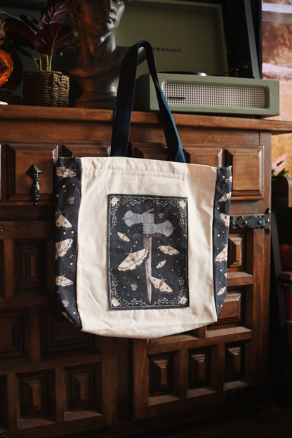The Knight and the Moth inspired tote bag with decorative design hanging on a wooden cabinet.