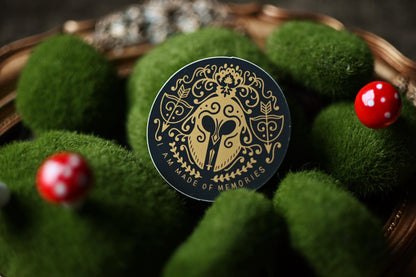 The matte version of the song of achillies inspired sticker by Madeline Mirror resting against a bed of fake moss. Showcases the golden detail design with the quote I am Made of Memories.