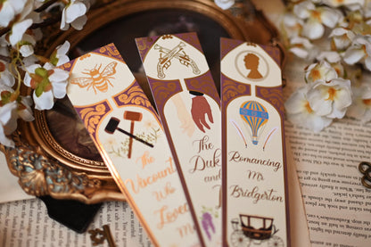 No One Designs Bridgerton-inspired bookmark collection with the three bookmarks fanned out against bookpages and a mirror that raised the bookmarks into the foreground. Each bookmark is inspired by one of Julia Quinn's books and features a gold-foil detailing. Bookmarks inspired by The Duke & I, The Viscount Who Loved Me and Romancing Mr Bridgerton. 