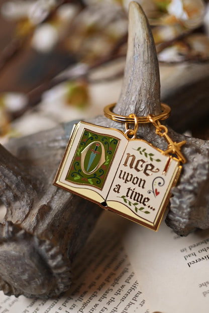 An enamel keychain with a gold metal frame and 'Once Upon a Time' text, displayed on a naturalistic setting with a leafy background.