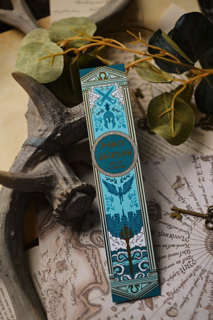 The Percy Jackson Book Spine inspired decorative bookmark with blue and gold design resting on a map with antlers and keys in the background