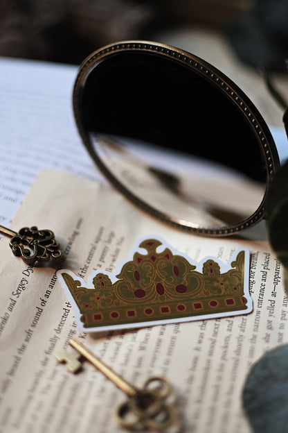 Inspired by V.E Schwab's A Darker Shade of Magic trilogy, Rhy's Golden Crown sticker rests against a backdrop of bookpages.