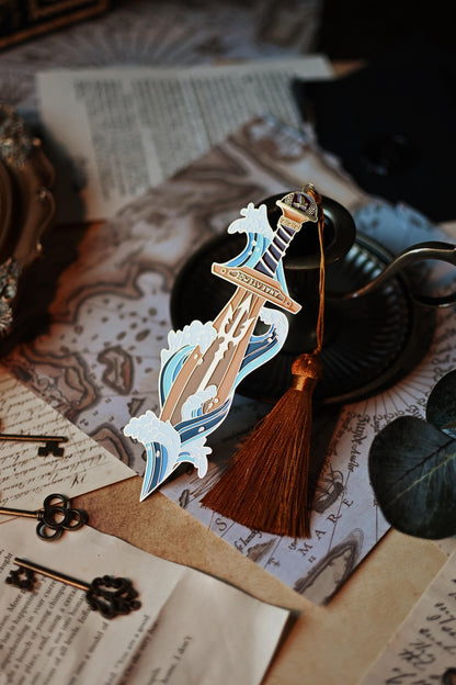 Percy Jackson inspired enamel pin featuring Percy's sword riptipe surounded by ocean waves. The bookmark is propped up against a candleholder and shines against the light to showcase a trident design in the sword. 