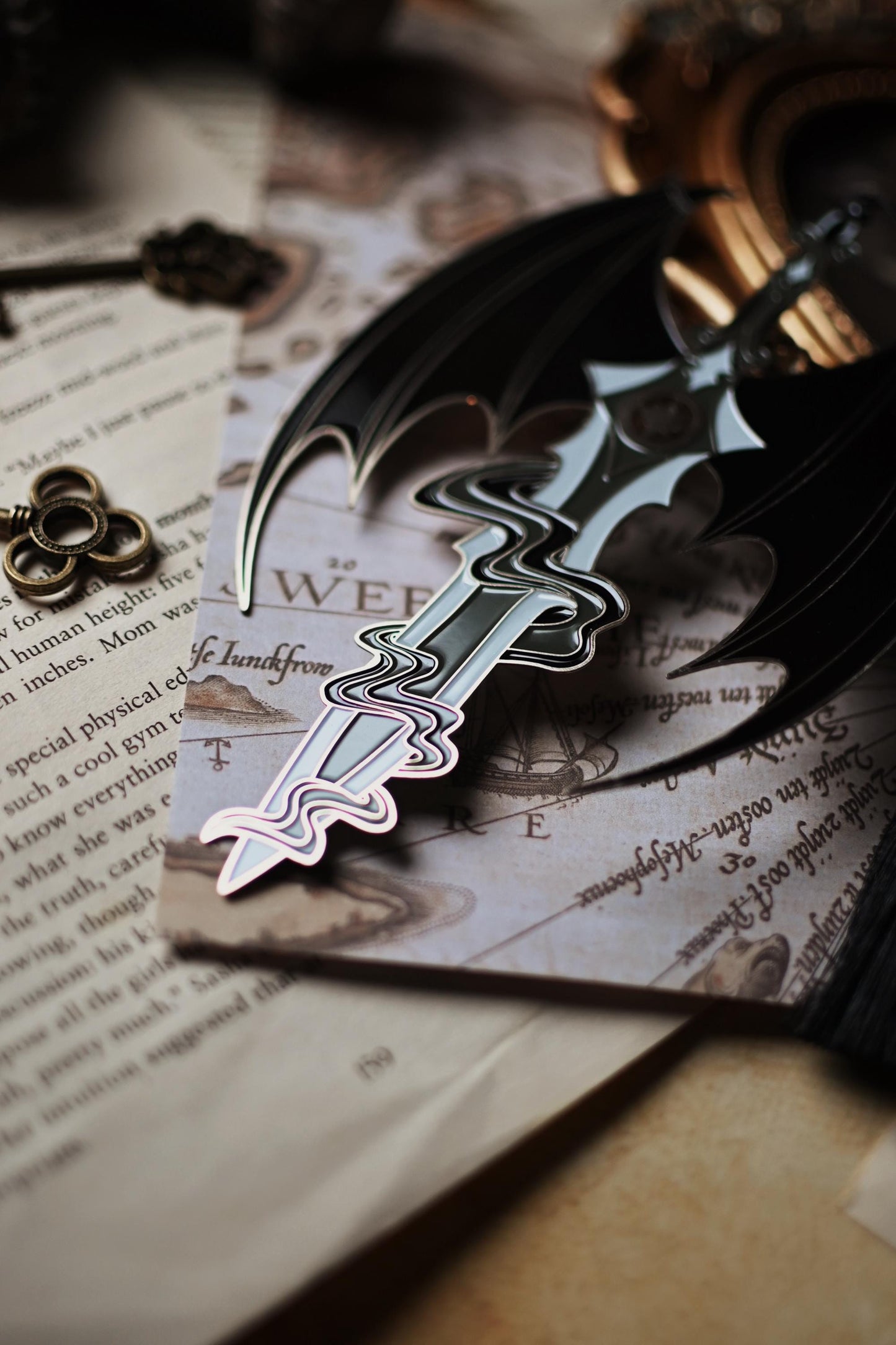 The officially licensed a court of thorns and roses bookmark inspired by the character of Azriel Shadowsinger. The enamel bookmark features a sword worthy of the night court and is surounded by the illyrian wings. The photo focuses on the tip of the sword which is surrounded in Azriel's shadows.