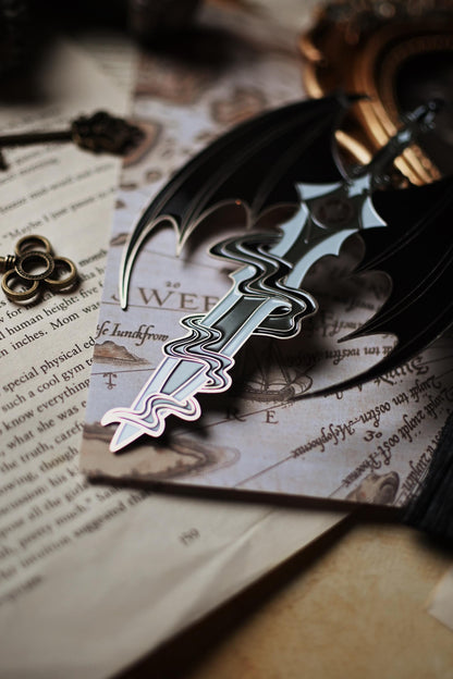 The officially licensed a court of thorns and roses bookmark inspired by the character of Azriel Shadowsinger. The enamel bookmark features a sword worthy of the night court and is surounded by the illyrian wings. The photo focuses on the tip of the sword which is surrounded in Azriel's shadows.