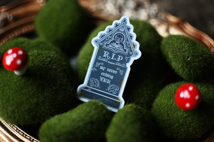 A silver foil decorative sticker with the text 'DEATH BY TBR' on a background that includes green moss and a wooden surface.