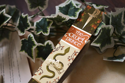 No One Designs book spine bookmark featuring a design inspired by The Cruel Prince by Holly Black. The bookmark is tilted up towards the camera to showcase the gold detailing and is resting against fake-vines. 