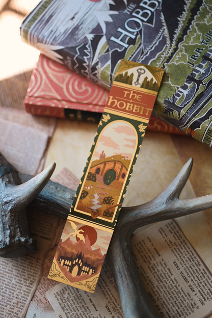 No0neDesigns foiled The Hobbit bookmark resting against copies of The Hobbit bby J.R.R Tolkien with the sunlight reflecting against the bookmark. In the background, there are book pages and the bookmark is being propped up against the book spines. 