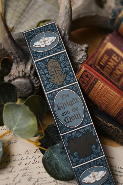 Decorative bookmark with 'The Knight and the Moth' design on a textured surface with books and antlers.