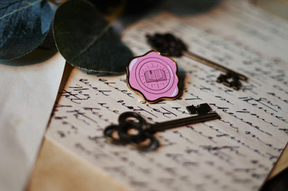 Enamel pin in the design of a wax seal with the centre featuring the outline of an open book. The pin rests against a background of written letters.