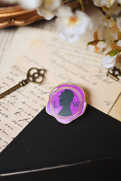 An enamel pin with a purple silhouette of a woman's profile, inspired by Lady Whistledown, placed on a black surface with a vintage background including a key and floral elements.
