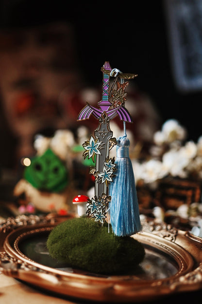 No0neDesigns Link's Master Sword enaml bookmark resting against the end of a painting to lift it against the bacground. The image shows the bookmark standing upright in a moss against the light with the background blurred but featuring a korok too. 