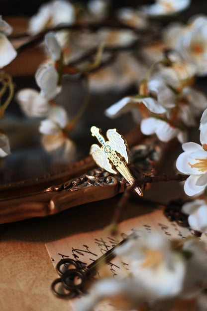 An enamel pin with a design resembling an angelic winged sword, placed among white flowers.