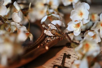 An enamel pin with a design resembling an angelic winged sword, placed among white flowers.