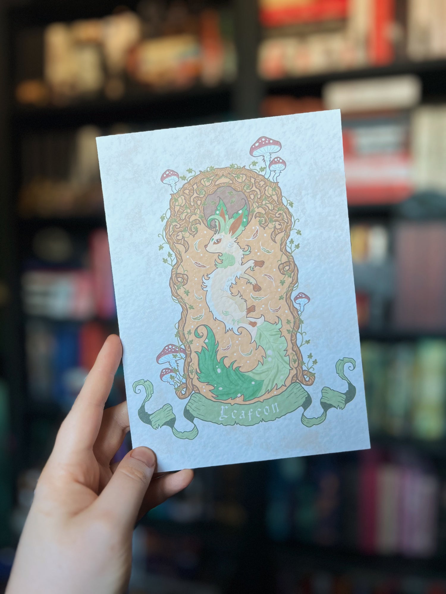 Chloe holding up one of the eevee inspired fox prints featuring a leafeon based fox surrounded by a boarder of roots with mushrooms sprouting out.